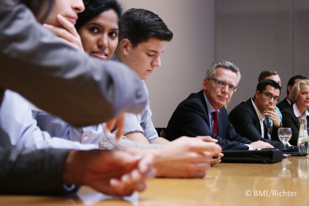 Foto von Thomas de Maizière, der an einem Tisch mit Teilnehmern sitzt.